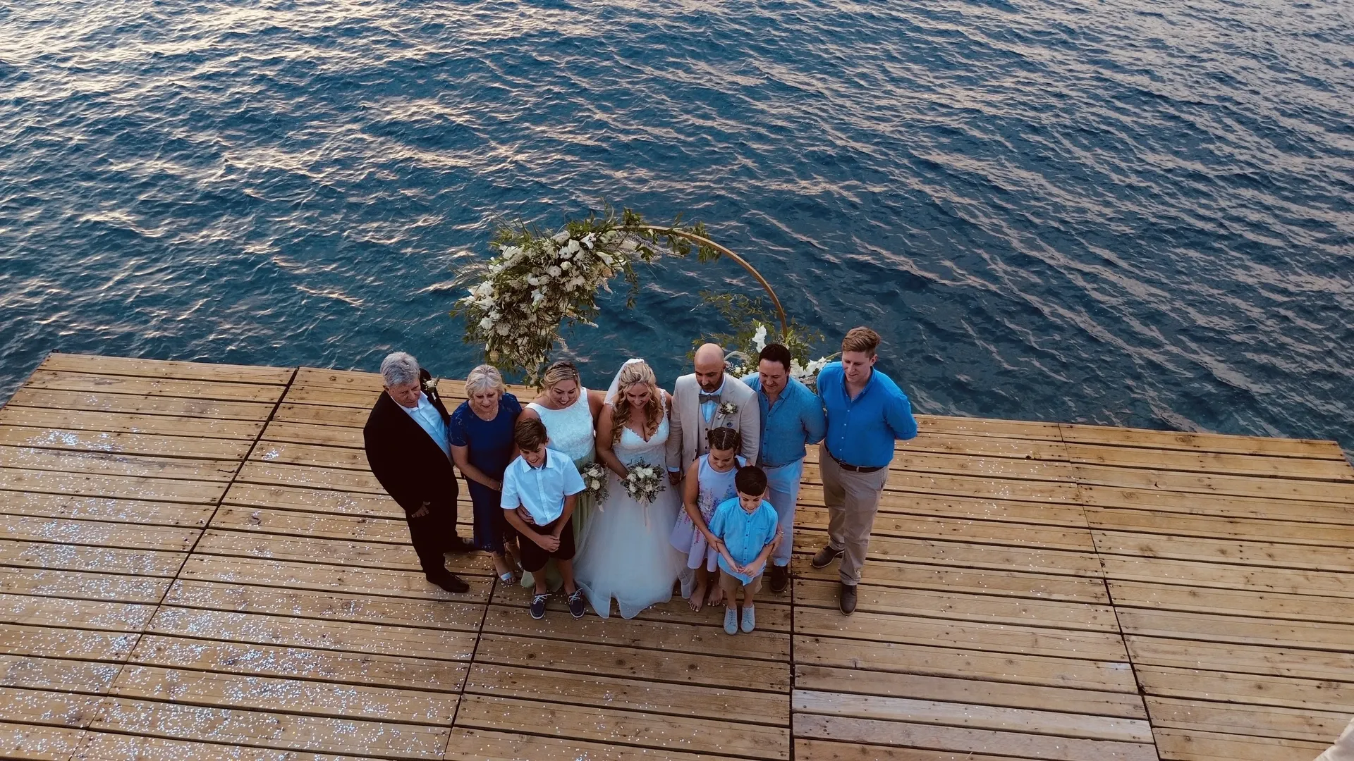 Drone view of wedding party gathered on wooden deck overlooking Mediterranean sea